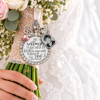A close-up of a bouquet wrapped in beige lace with a silver-toned keepsake charm featuring a wedding message, a small portrait photo, and a pink pearl accent, held by a hand against a white lace dress background.