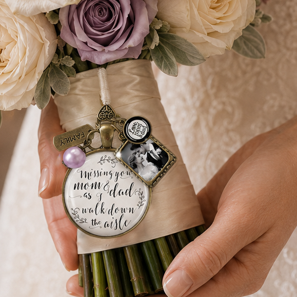 A close-up of a bouquet charm with a black and white photo of parents, hanging on a bridal bouquet of cream and lavender roses with green foliage, held by a bride  in a white lace wedding dress. The bouquet is wrapped in beige ribbon with a round glass pendant that says "Missing you mom & dad as I walk down the aisle? and meaningful decorative charms.