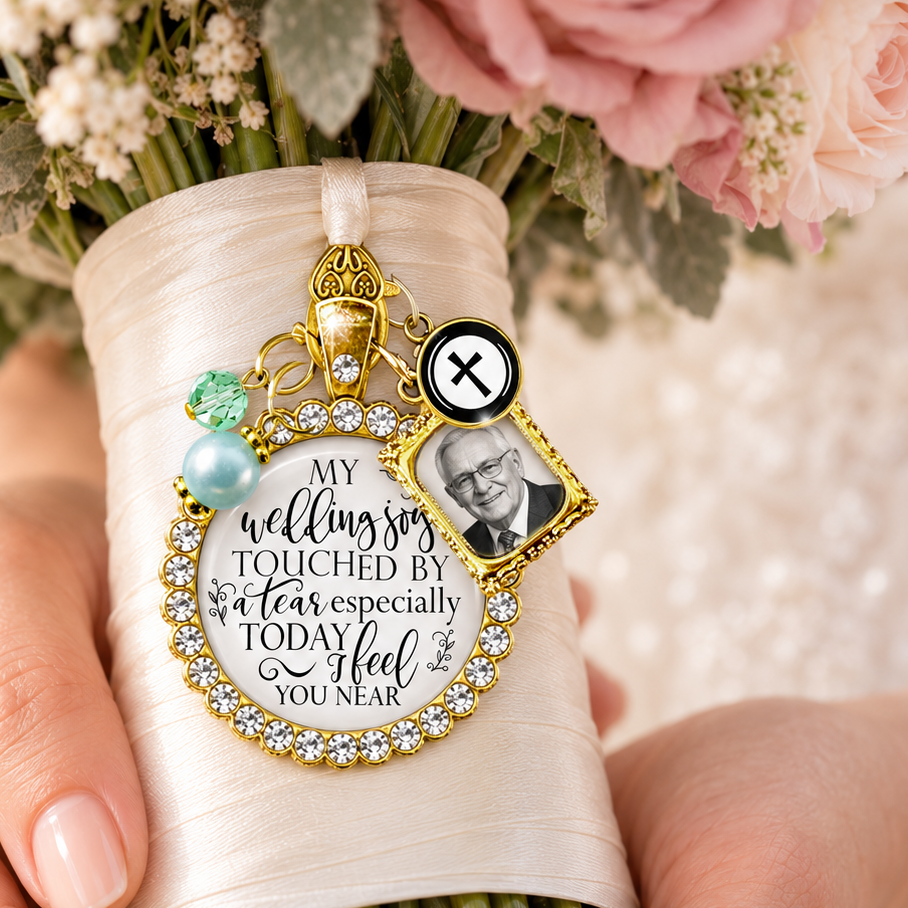 Custom gold bridal bouquet memorial charm with rhinestone border, personalized photo frame, crystal cross charm, and sentimental quote pendant pinned to a cream satin ribbon-wrapped wedding bouquet with dusty rose flowers.