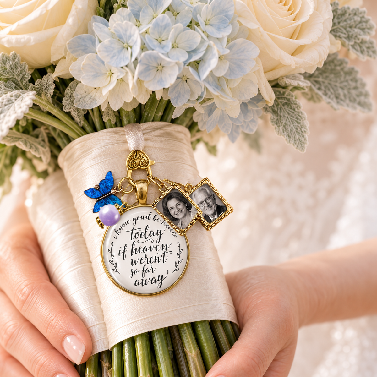 Bride holding cream and blue hydrangea wedding bouquet wrapped in satin ribbon adorned with a custom gold memorial charm featuring two personalized photo frames, blue butterfly charm, lavender pearl, and scripted quote: "I know you'd be here today if heaven weren't so far away."