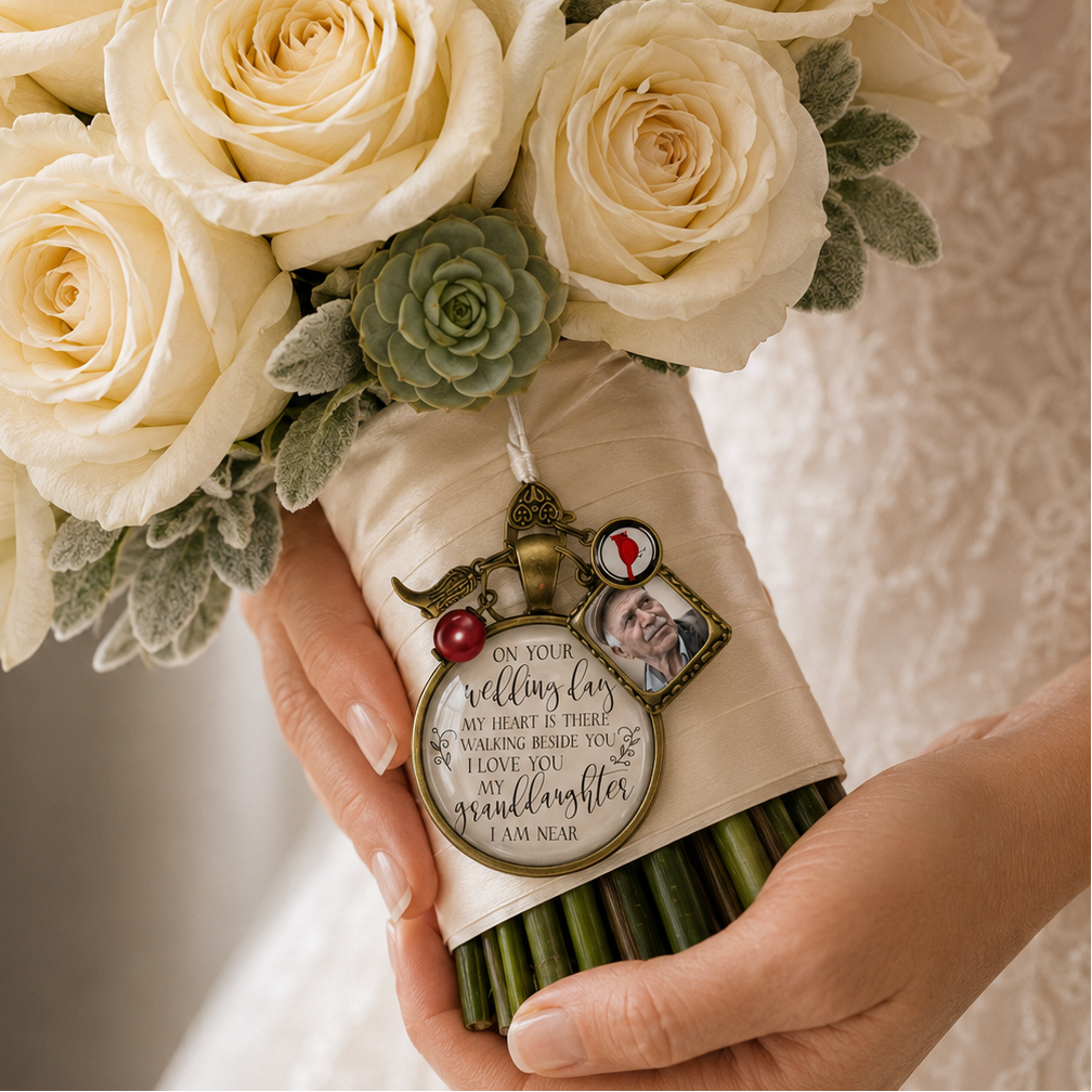 Bride holding cream rose wedding bouquet with a personalized bronze memorial charm featuring a grandfather photo frame, red cardinal charm, ruby pearl bead, angel wing, and scripted quote: "On your wedding day my heart is there, walking beside you I love you my granddaughter I am near."
