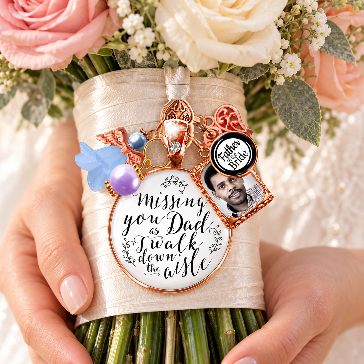 Bride holding ivory and pink rose wedding bouquet wrapped in satin ribbon adorned with a custom rose gold memorial charm featuring a father photo frame, "Father of the Bride" charm, something blue angel wing, purple pearl, and scripted quote: "Missing you Dad as I walk down the aisle."