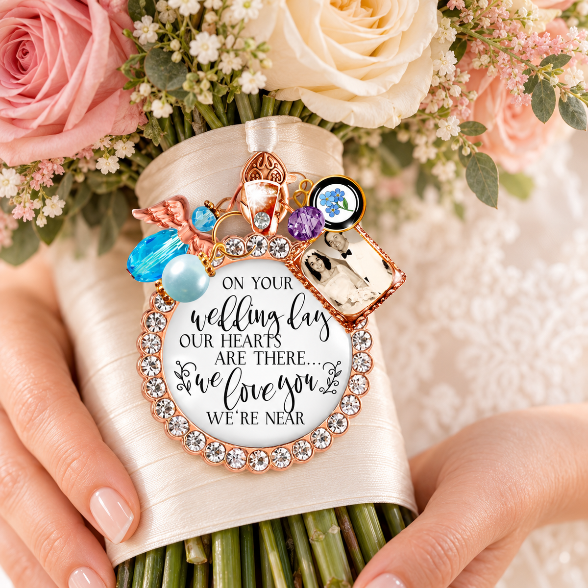 Bride holding pink rose wedding bouquet wrapped in cream satin ribbon with a custom rose gold memorial charm featuring a sparkling rhinestone border, personalized photo frame, crystal cross, blue crystal, and scripted quote: "On your wedding day our hearts are there... we love you we're near."