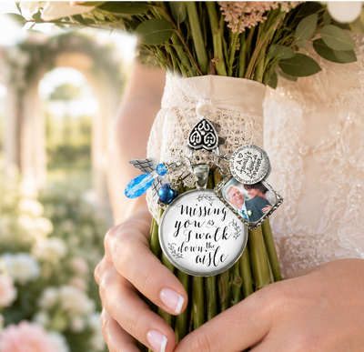 A close-up of a bouquet wrapped in a cream lace wristband, held by a person in a white lace dress. The bouquet is decorated with silver charms, including a round pendant that reads 'Missing you? I walk down the aisle.